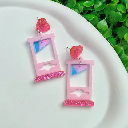 Kiss Me Later’s sparkly acrylic earrings feature pink window shapes with blue and white panes, heart cutouts, glittery pink bases, and red glitter heart studs, displayed on a white surface with green leaves in the background.