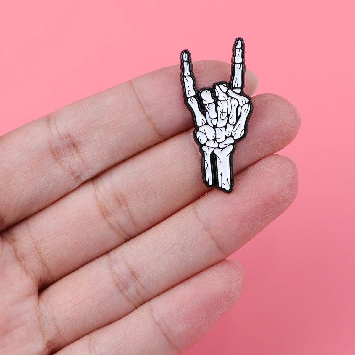 A hand holds the Kiss Me Later skeleton hand enamel pin making the rock on gesture, adding spooky vibes against a pink background.