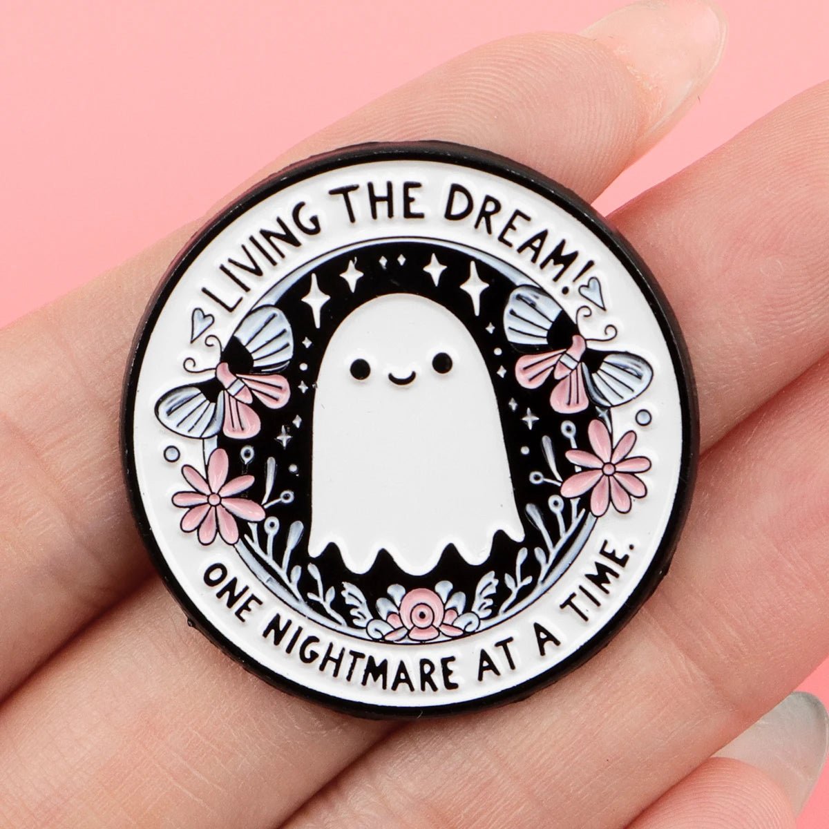 A round ghost pin, shown in hand, features an adorable ghost with flowers and butterflies. The edge reads LIVING THE DREAM! ONE NIGHTMARE AT A TIME. Black background is dotted with stars.