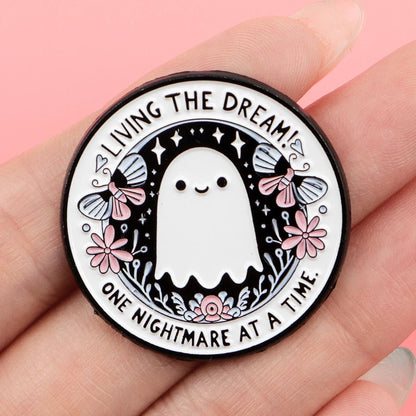 A round ghost pin, shown in hand, features an adorable ghost with flowers and butterflies. The edge reads LIVING THE DREAM! ONE NIGHTMARE AT A TIME. Black background is dotted with stars.
