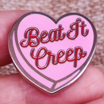 A hand holds an enamel pin shaped like a love heart in pink and red, featuring bold red text that says, Beat It Creep.