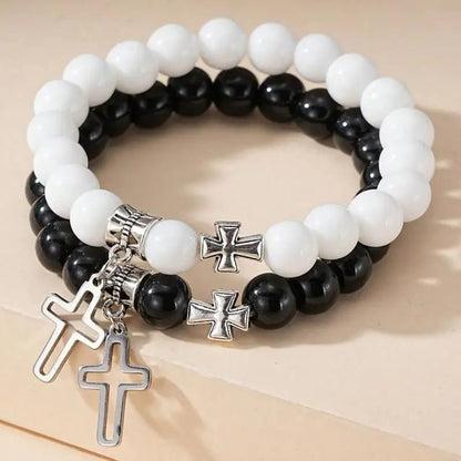 Two beaded crystal bracelets by Kiss Me Later, with white and black round beads, each featuring a silver cross charm and a cross-shaped bead, displayed on a light beige surface.