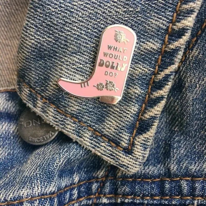 A pink and gold cowboy boot pin with What would Dolly do? and floral accents is the perfect addition to any Dolly-themed enamel pin collection, shown here on a denim jacket.
