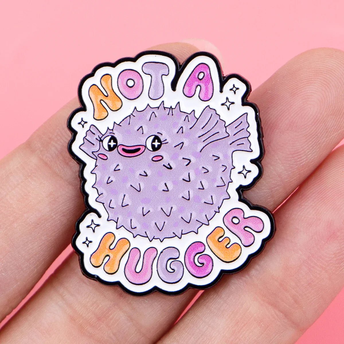 A hand holds a cute purple pufferfish pin by Gay Skate, featuring a big smile and Not A Hugger in colorful bubble letters—a playful pride accessory—set against a pink background.