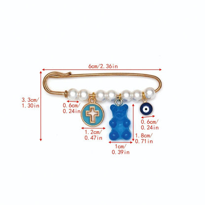 The Kiss Me Later gold safety pin features five white beads and three dangling charms—a round cross, a blue gummy bear, and a blue evil eye—making it a unique evil eye pin. Dimensions are listed in centimeters and inches.