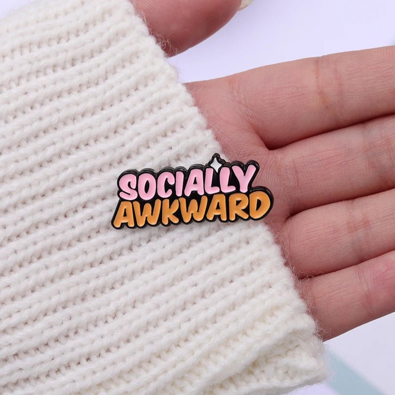 A hand in a white knitted sleeve holds an enamel pin featuring the phrase SOCIALLY AWKWARD in bold pink and orange letters.