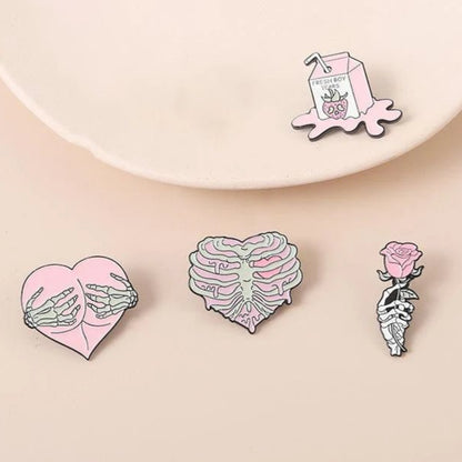 Four pastel enamel pins: a skeleton with a pink rose, skeletal hands holding a pink heart, a rib cage forming a heart, and a cute pink milk carton pin with a crying face and straw.