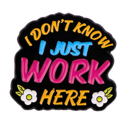 Enamel pin features I DONT KNOW I JUST WORK HERE in colorful letters on a black background with two white flowers—an ideal fun coworker gift or emotional support pin for the office.