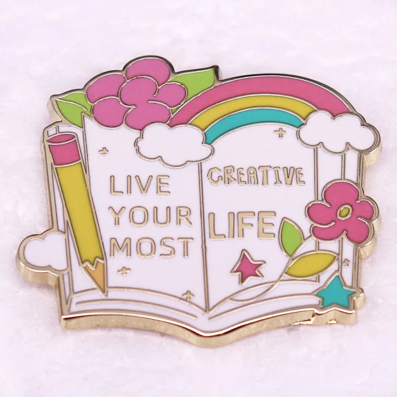 Rainbow pin for book lovers features an open book with flowers, a rainbow, clouds, pencil, and stars. “LIVE YOUR MOST CREATIVE LIFE” is printed in gold on white—perfect for any Romance Reader.