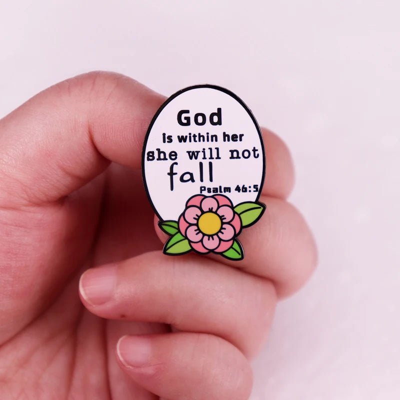 A hand holds a white oval enamel pin with a pink flower at the bottom. The pin reads: God is within her she will not fall. Psalm 46:5.