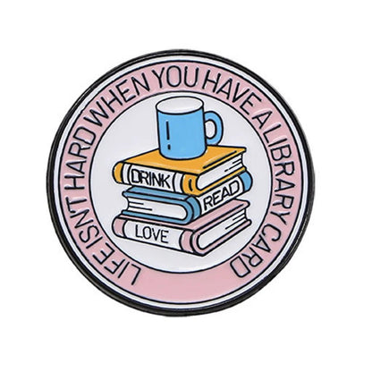 Round enamel pin features a blue mug atop three stacked books labeled DRINK, READ, and LOVE. The rim says LIFE ISNT HARD WHEN YOU HAVE A LIBRARY CARD—a perfect gift for book or romance lovers.