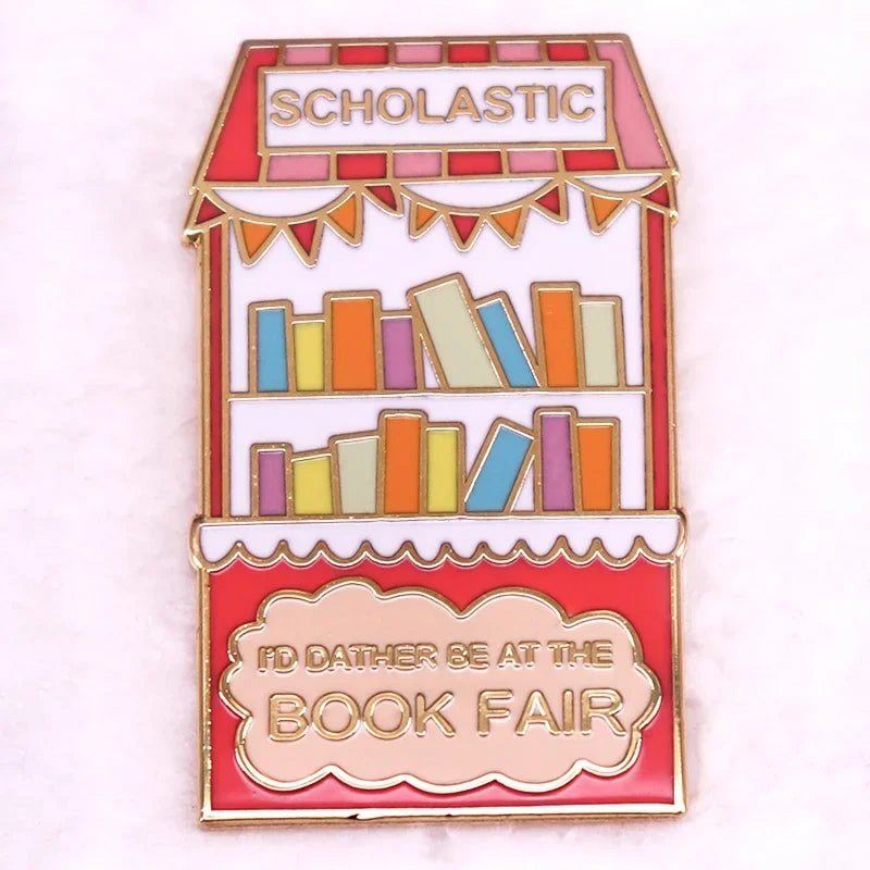 Enamel pin shaped like a red book fair stand, decorated with colorful books and bunting. Features SCHOLASTIC on top and ID RATHER BE AT THE BOOK FAIR in gold—perfect for book lovers!.