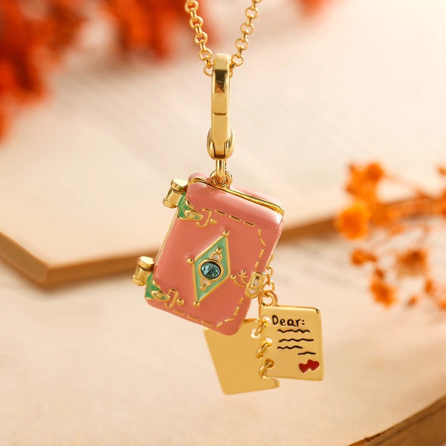Gold pink enamel pendant, shaped like an opening book with a blue gem, green accents, and a love letter charm reading Dear: with a red heart, rests on a warm background adorned with orange flowers.