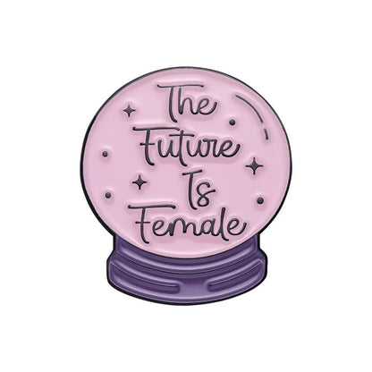 An enamel pin shaped like a pink crystal ball with black “The Future Is Female” text and tiny stars, sitting on a purple heart-shaped base, shown against a white background.