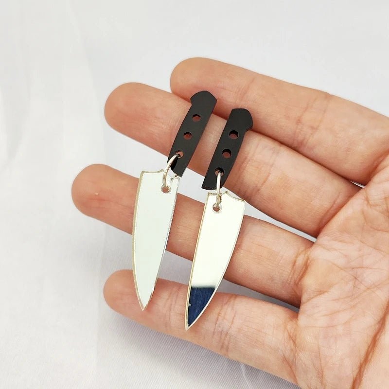 A hand holds a pair of Kiss Me Later knife earrings with shiny silver acrylic blades and black handles, shown against a white background.