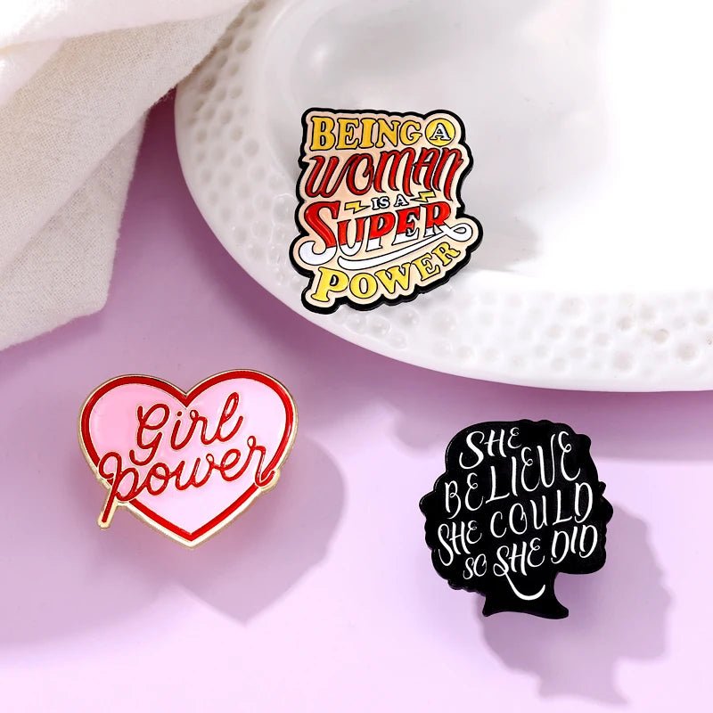 Three enamel pins on a pink surface: a heart with Girl power, one with Being a woman is a super power, and a woman’s head with She believed she could so she did—perfect for celebrating self love.