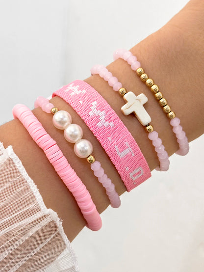 A wrist adorned with a Kiss Me Later pink bracelet stack—five pink and gold beaded bracelets, including a woven white-letter bracelet and a WWJD design. The person is wearing a white mesh sleeve.