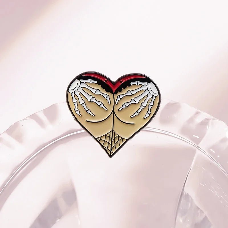 Enamel pin features a heart shape with beige and red Gothic spiderweb designs at the bottom and skeleton hands reaching down from the top, displayed on a clear, textured surface.