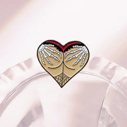 Enamel pin features a heart shape with beige and red Gothic spiderweb designs at the bottom and skeleton hands reaching down from the top, displayed on a clear, textured surface.