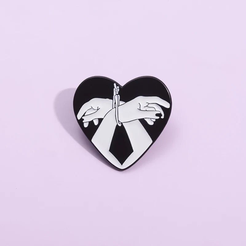 The Kiss Me Later enamel pin features two hands handcuffed together in a love heart shape on a light purple background—a bold black and white accessory for striking self-expression.