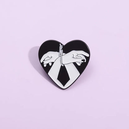 The Kiss Me Later enamel pin features two hands handcuffed together in a love heart shape on a light purple background—a bold black and white accessory for striking self-expression.