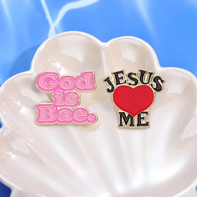 Two decorative Kiss Me Later enamel pins on a white seashell dish: one is a cute pink “God Is Bae” pin; the other features “Jesus [heart] Me” with a red heart on a blue background.