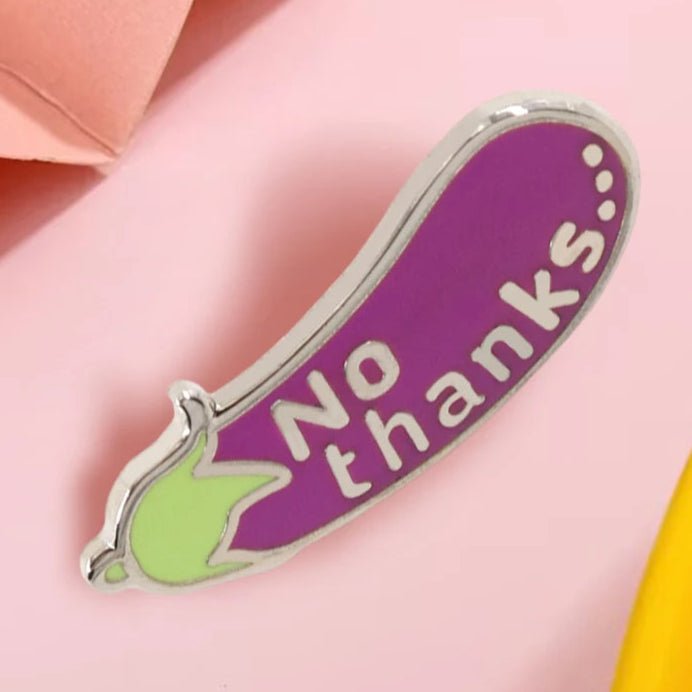 Enamel pin is shaped like a purple eggplant with a green top and the words No thanks in white, shown on a pink surface.