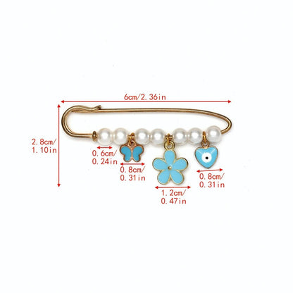 The Kiss Me Later gold safety pin features five pearls and three blue charms—a butterfly, flower, and heart—making it a sweet baby shower or evil eye gift. Red measurements label its beads and charms in centimeters and inches.