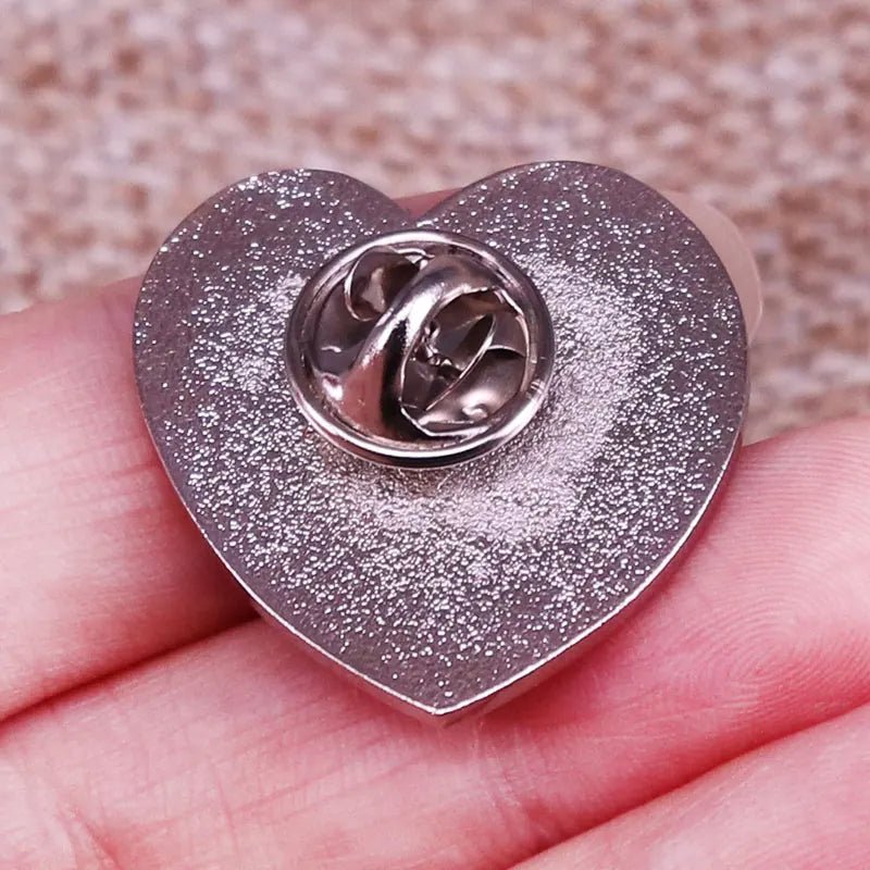 A hand holds a silver love heart enamel pin with a butterfly clutch fastener, displayed against a textured background.