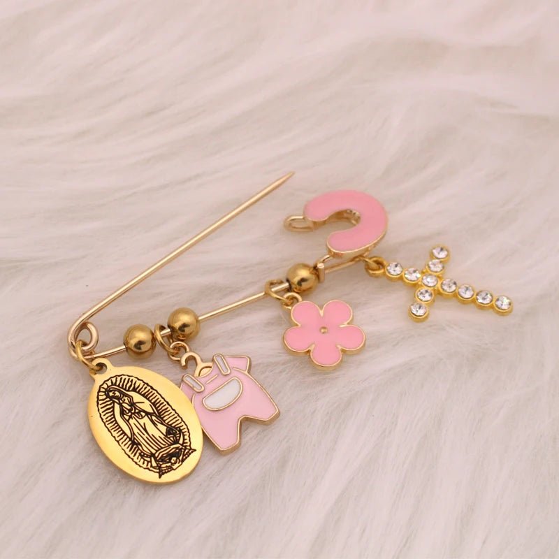 A Kiss Me Later gold baby pin brooch features four charms: an oval Virgin Mary medallion, a pink Among Us figure, a pink flower, and a sparkling diamante cross. The pin is displayed on soft white fur.