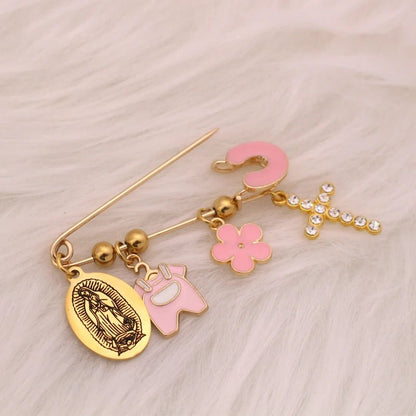 A Kiss Me Later gold baby pin brooch features four charms: an oval Virgin Mary medallion, a pink Among Us figure, a pink flower, and a sparkling diamante cross. The pin is displayed on soft white fur.