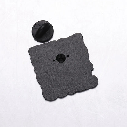 A black, scallop-edged square Jesus pin with a central fastener and two small holes sits on a white surface. The round black pin back is detached and placed above the pin.