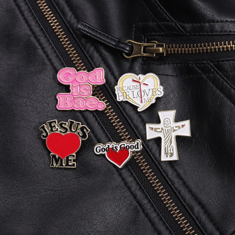 Five Kiss Me Later religious enamel pins, like the pink God Is Bae pin, are displayed on a black leather jacket with a zipper. Designs include “Jesus ♥ Me” and heart-accented crosses.