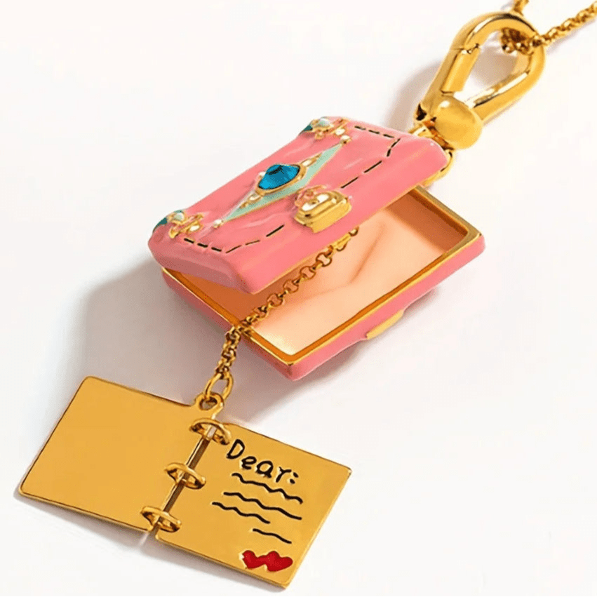 My Cutesy Eras gold necklace showcases a pink enamel book-shaped envelope pendant with a blue gem. It opens to reveal a gold, book-shaped charm engraved with Dear: and red heart accents.