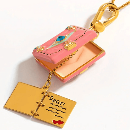 My Cutesy Eras gold necklace showcases a pink enamel book-shaped envelope pendant with a blue gem. It opens to reveal a gold, book-shaped charm engraved with Dear: and red heart accents.