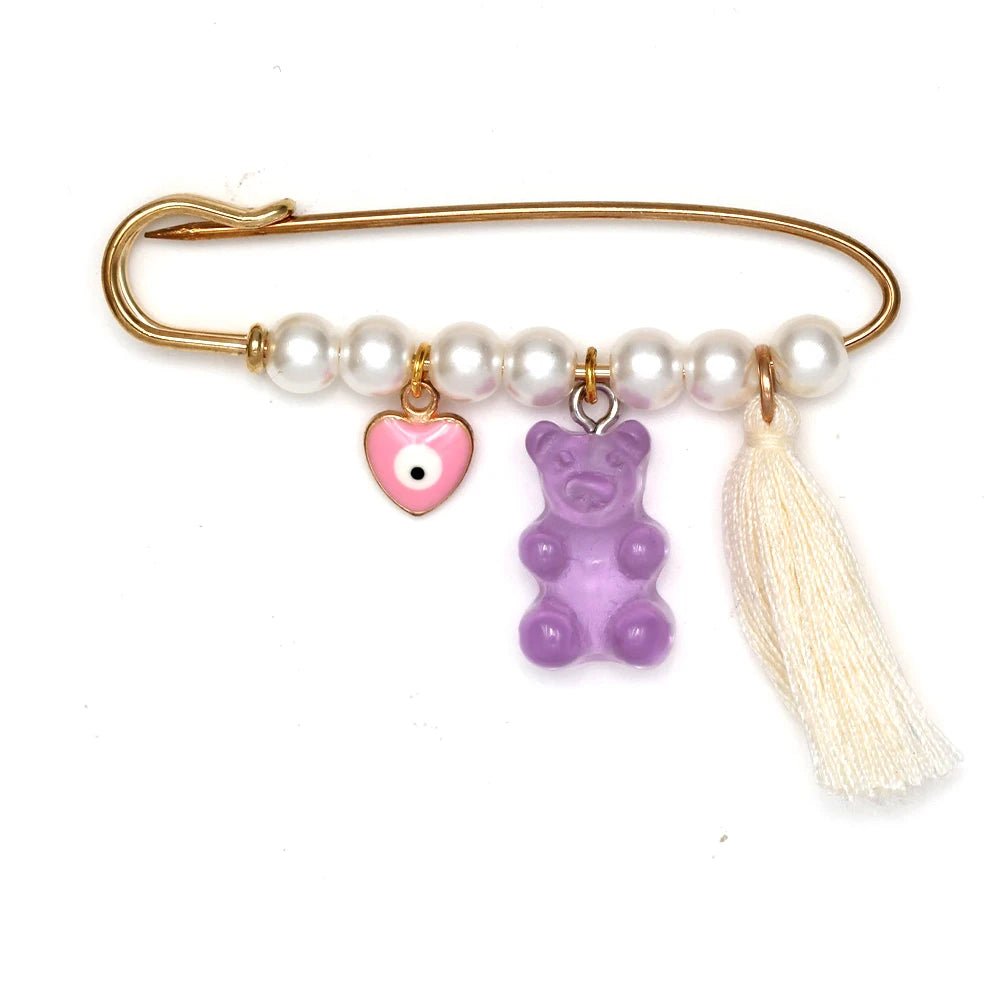 A gold safety pin brooch by Kiss Me Later features white pearls, a pink heart evil eye pin charm, a purple gummy bear charm, and a white tassel—making it a perfect baby shower gift.
