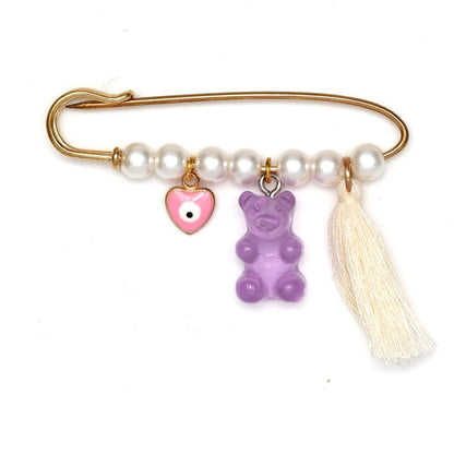A gold safety pin brooch by Kiss Me Later features white pearls, a pink heart evil eye pin charm, a purple gummy bear charm, and a white tassel—making it a perfect baby shower gift.