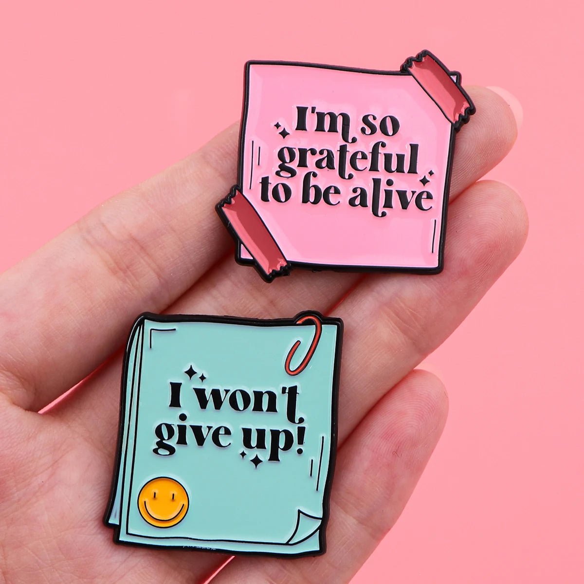 A hand holds two colorful pins with affirmations—“I’m so grateful to be alive” and “I won’t give up!”—promoting self empowerment and mental health on a cheerful light pink background.