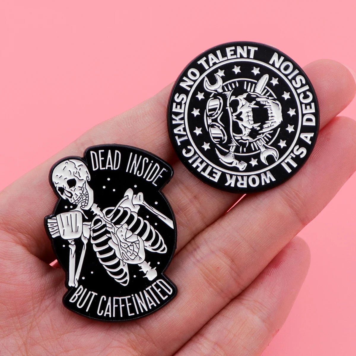 A hand holds two Kiss Me Later skeleton hand enamel pins on a pink background—one says Dead Inside But Caffeinated, the other reads Work Ethic Takes No Talent, It’s A Decision.