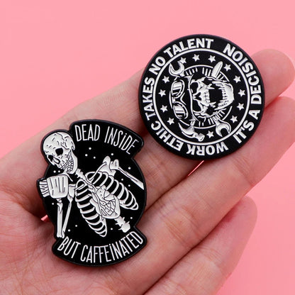 A hand holds two Kiss Me Later skeleton hand enamel pins on a pink background—one says Dead Inside But Caffeinated, the other reads Work Ethic Takes No Talent, It’s A Decision.