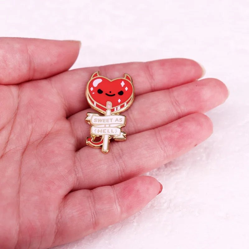 A hand holds a Kiss Me Later enamel pin shaped like a smiling red devil heart with tiny horns, tail, and a banner that says SWEET AS HELL.