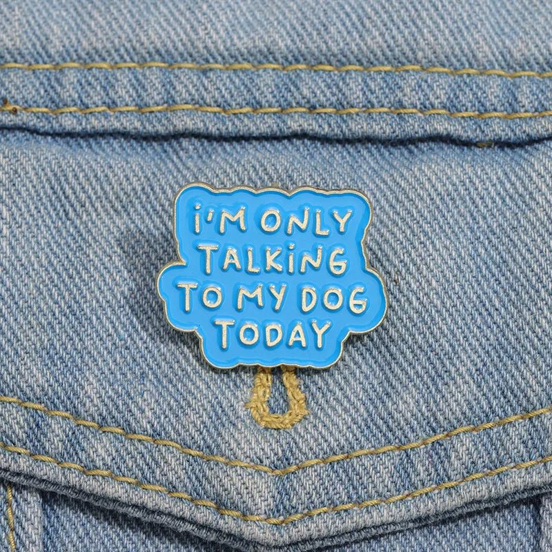 A blue enamel pin by featuring white text that says IM ONLY TALKING TO MY DOG TODAY, is pinned to the pocket of a light blue denim jacket—an ideal accessory for proud dog lovers.