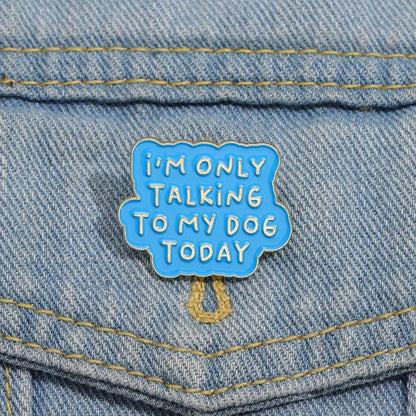 A blue enamel pin by featuring white text that says IM ONLY TALKING TO MY DOG TODAY, is pinned to the pocket of a light blue denim jacket—an ideal accessory for proud dog lovers.