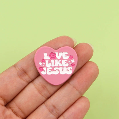 A hand holds a Kiss Me Later pink heart pin with white Love Like Jesus text and small pink flowers, set against a light green background.