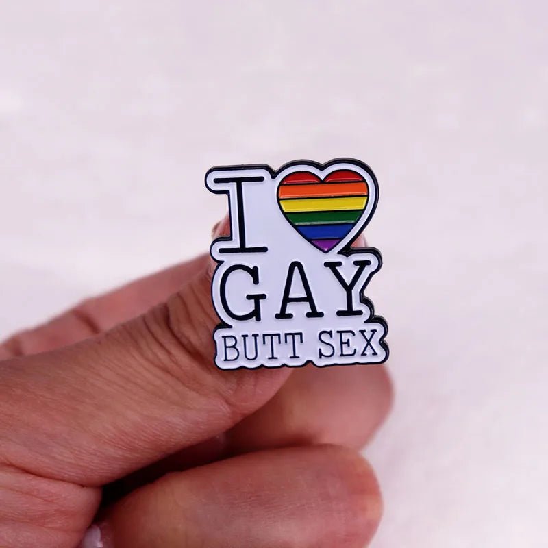 A hand holds a Kiss Me Later enamel pin that reads I ❤ GAY BUTT SEX, with the heart in rainbow stripes to represent LGBTQ+ pride.