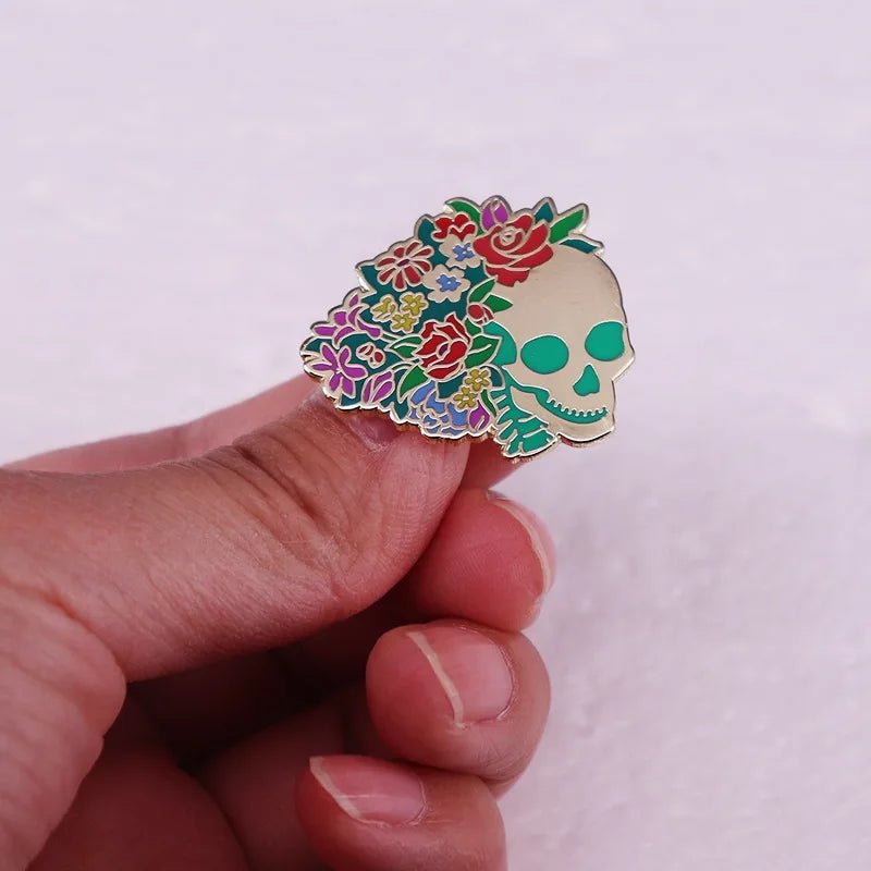 A hand holds an enamel pin featuring a skull adorned with a vibrant Gothic bouquet of colorful flowers and leaves on one side, set against a light textured background.