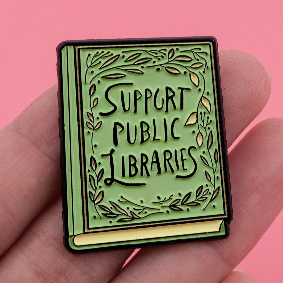 A hand holds a green enamel book-shaped pin featuring the message Support Public Libraries—a perfect accessory for any booktrovert, shown against a pink background.