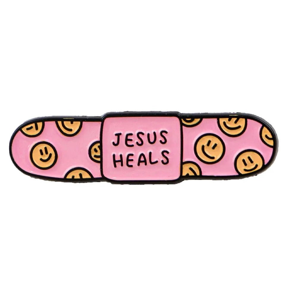 pink bandaid enamel pin with yellow smiley faces on the ends and JESUS HEALS printed across the center.