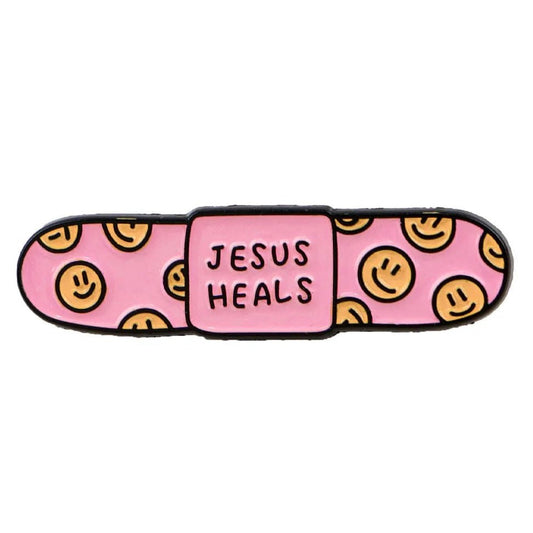 pink bandaid enamel pin with yellow smiley faces on the ends and JESUS HEALS printed across the center.