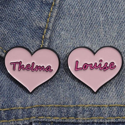 Two pink heart-shaped enamel pins by My Cutesy Era, featuring Thelma and Louise in pink script, are attached to blue denim fabric—celebrating iconic feminism and strong women.